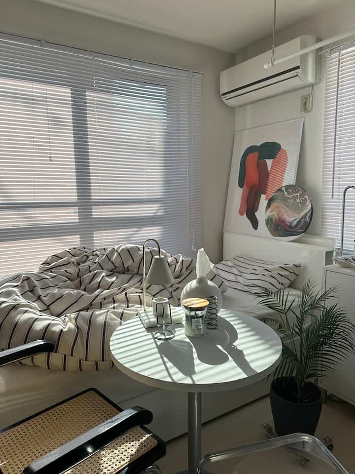 Bright white tone bedroom featuring a neat bed, blinds, a small round table, and a potted plant in a cozy setting