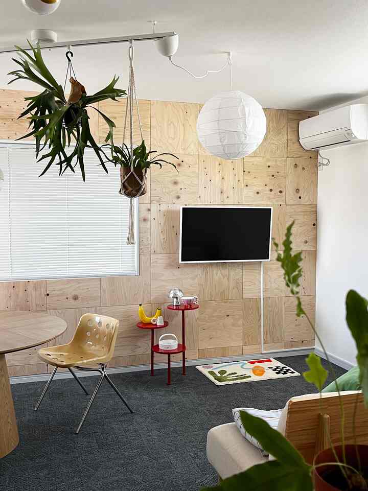 白い壁と木目調のパネルが調和したリビング、ベージュのダイニングチェアと赤いサイドテーブル、緑の植物が配置された雰囲気のある空間