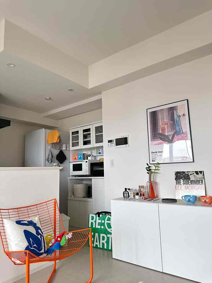 明るいホワイトトーンのリビングとキッチンが繋がる空間、オレンジのワイヤーチェアとすっきりした収納キャビネットが配置されたモダンなインテリア