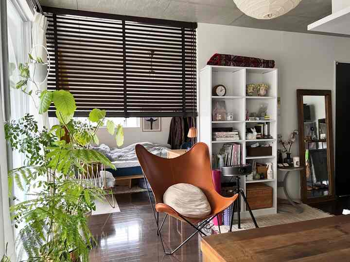 Natural and modern single household living bedroom featuring brown butterfly chair, dark brown blinds, wooden floor, and abundant green plants