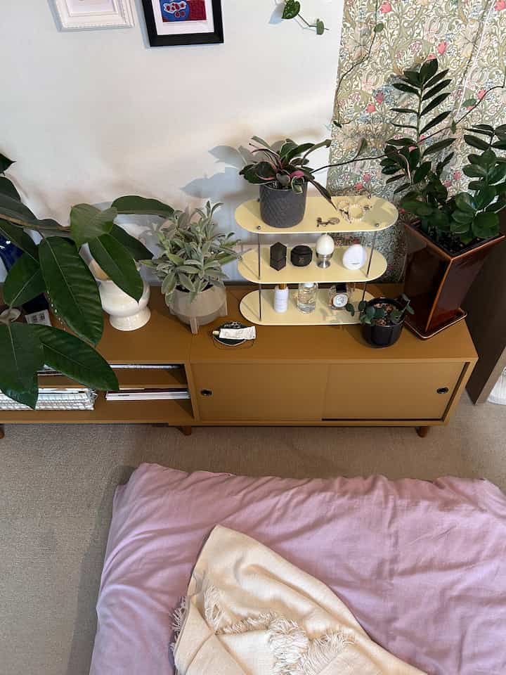 ナチュラルトーンのリビング、マスタード色のチェストの上に多種多様な植物とラベンダー色の寝具が配置された暖かみのあるインテリア