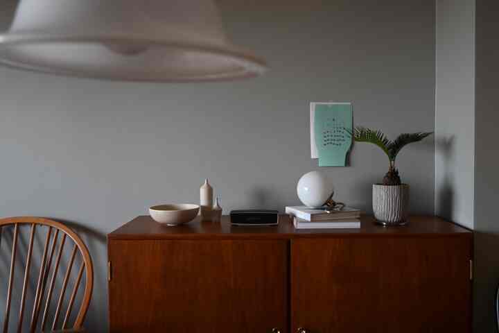 グレーの壁と木目調のチェスト、植物とテーブルランプが配置されたカフェ風のナチュラルなリビングスペース