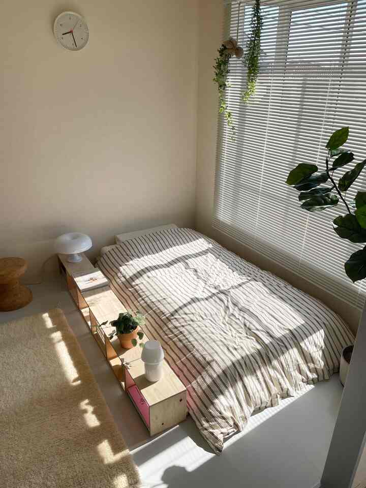 明るく自然な色調の寝室、中央にストライプ柄の掛け布団カバーが敷かれたベッドと窓際の植物が配置された落ち着いたインテリア