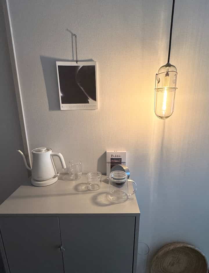 モノトーンのキッチンスペース、白いキャビネットの上に電気ケトルとガラス容器が置かれ、暖かい間接照明が雰囲気を演出している