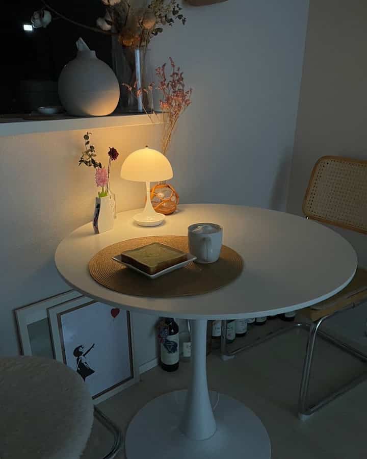 ナチュラルな色調のリビング、円形の白いローテーブルにテーブルランプと朝食が置かれたシンプルで温かみのある雰囲気のインテリア