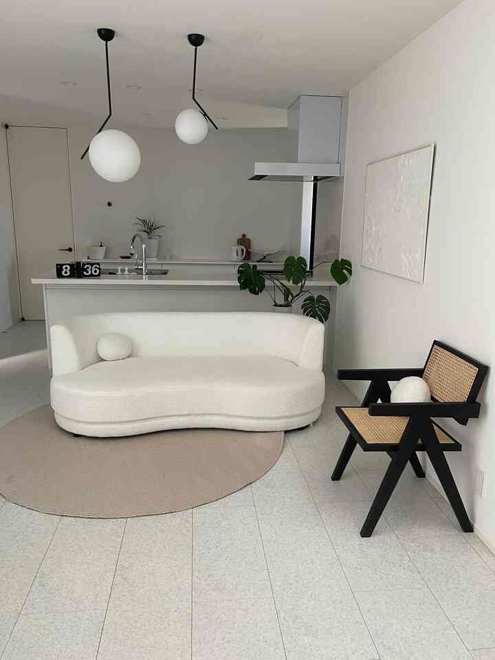 A modern living room and kitchen with white and beige tones featuring a simple curved sofa and a rattan armchair