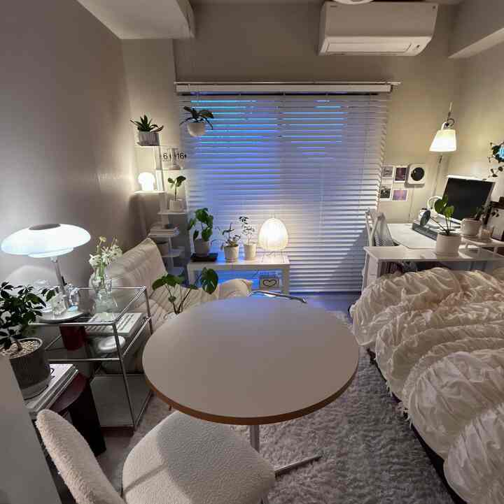 White-toned compact studio apartment featuring a round dining table, sofa on left, bed on right, and white blinds creating a cozy atmosphere