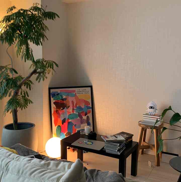 ナチュラルな色調のリビング、ブラックのテーブルと木製スツール、大きな植物と暖かな照明が配置された居心地の良い空間