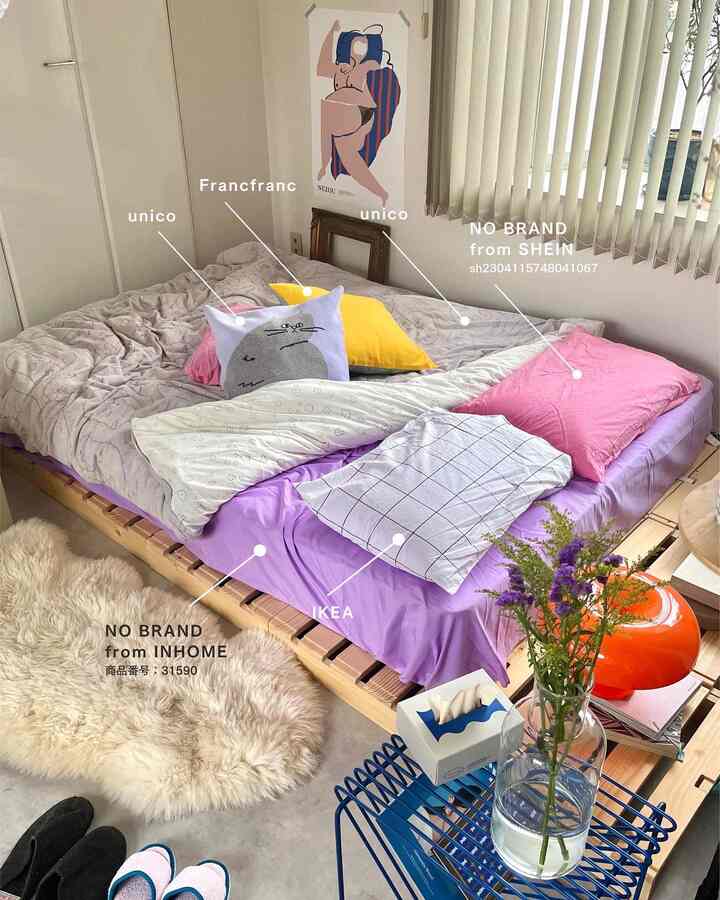 ラベンダーとピンクの枕が置かれた木目調のベッドとポスターがある狭い寝室