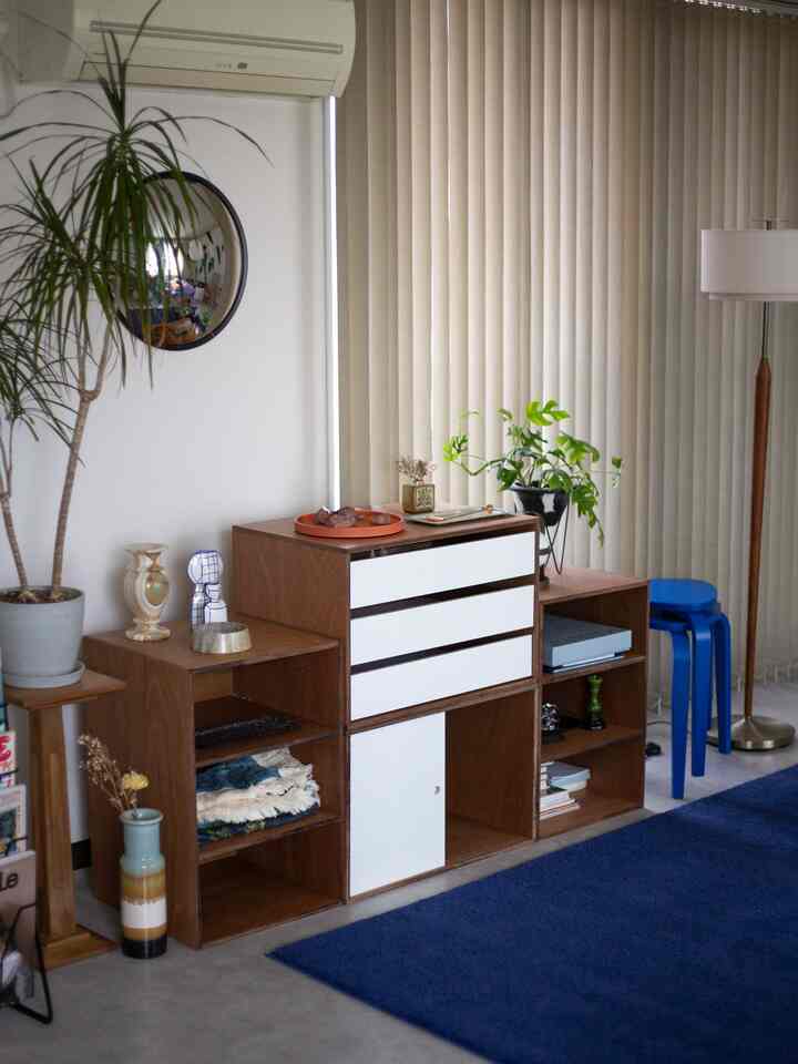 ブラウンとホワイトのリビング収納棚、ブルーのラグ、縦型ブラインドが配置された落ち着いた空間