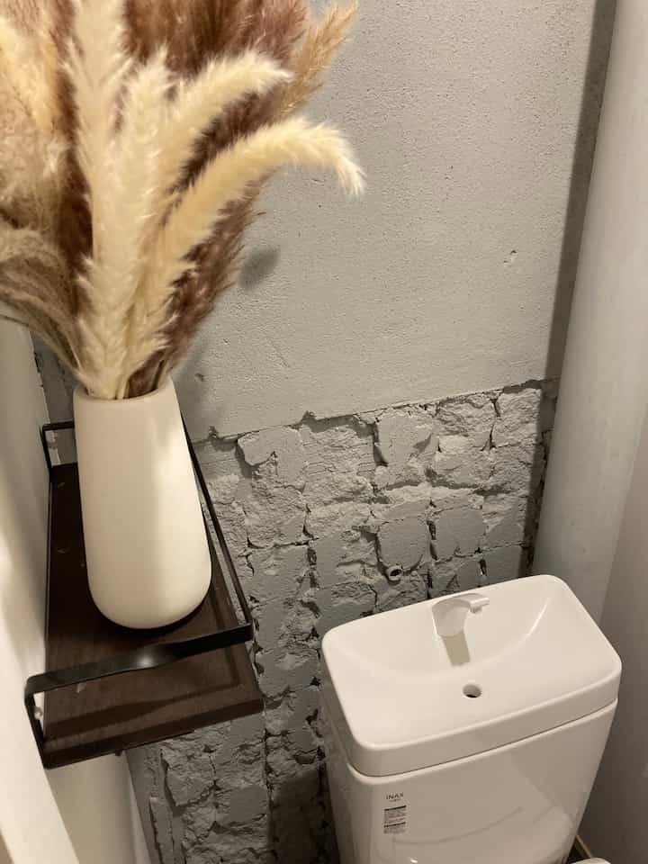 ナチュラルカラーのトイレ、ベージュのパンパスグラスブーケとダークウッドの棚が配されたテクスチャーのあるグレー石壁とシンプルな便器のある雰囲気のあるインテリア