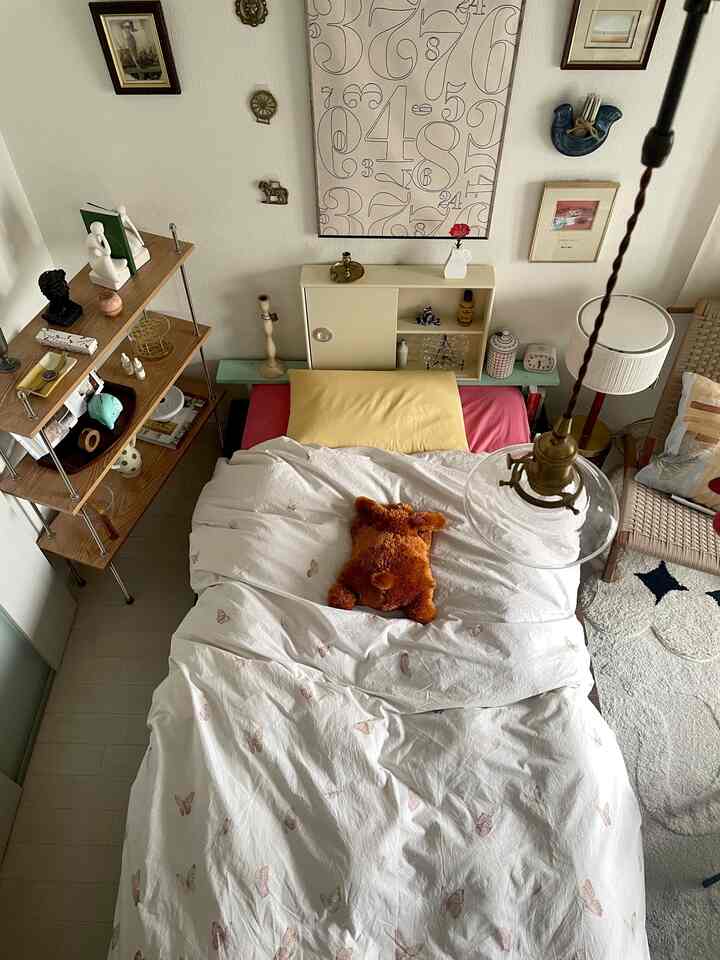 Cozy bedroom with white and wood tones, featuring a bed with a yellow pillow and plush toy centrally arranged, complemented by a wooden shelf in a natural setting