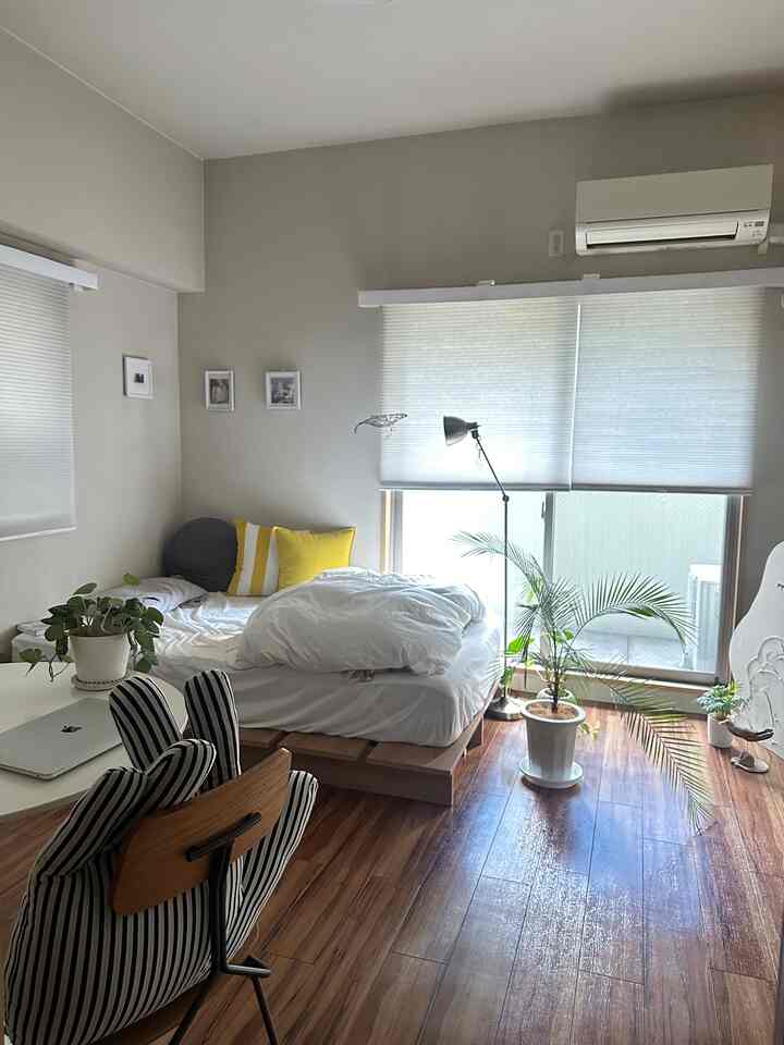 白と木目調が調和した一人用寝室、広い窓のブラインドとフロアランプ、植物が配置された雰囲気のある空間