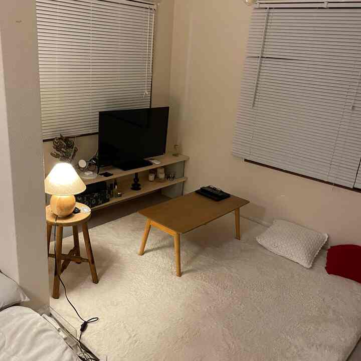 ナチュラルなウッドトーンとホワイトの色調が調和したコンパクトなワンルームのリビング兼寝室空間。ミニマルな家具配置とテーブルランプが印象的なシンプルなインテリア