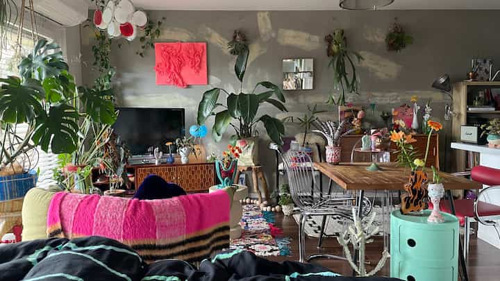 グレーの壁に多彩な植物が配置された狭いリビング、木製のダイニングテーブルと鮮やかなピンクの毛布が印象的なナチュラルなインテリア