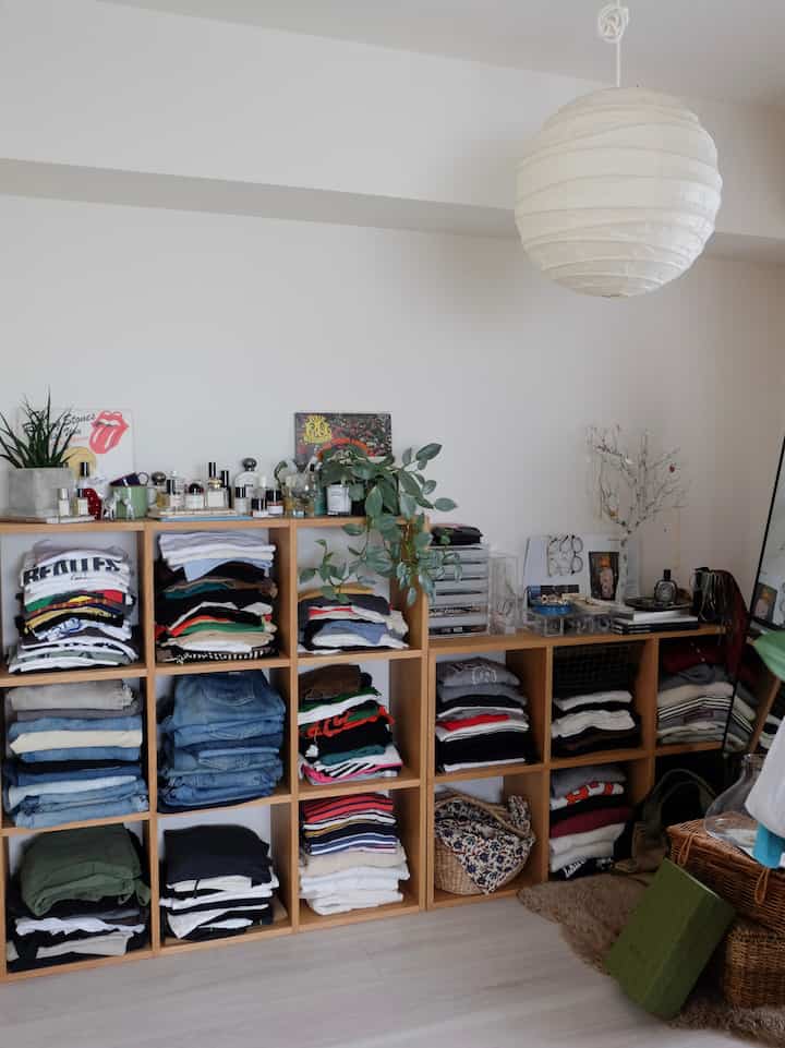 明るく自然な色調のクローゼット空間、折りたたみ式の木製棚に整理された衣服と植物が配置された清潔感のあるインテリア