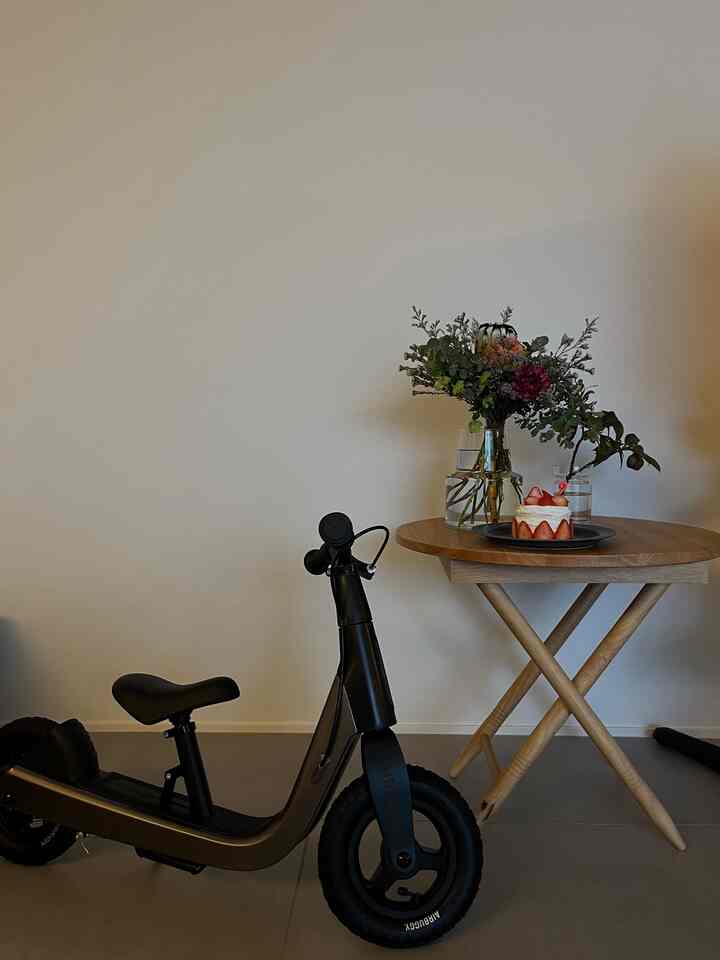 ウッドトーンの丸テーブルにイチゴのケーキと花が置かれた子供部屋、黒い電動キックボードが配置されたシンプルな室内