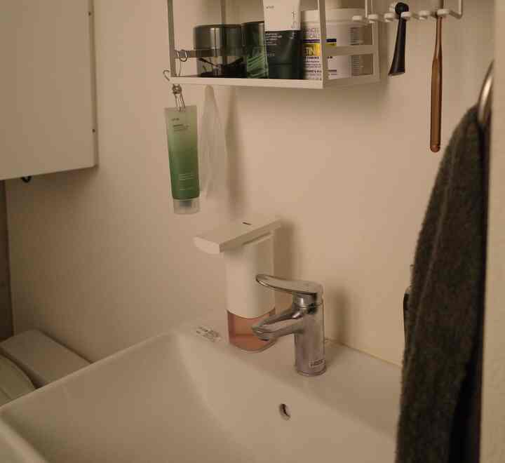 White-toned bathroom sink featuring an automatic foam dispenser and wall shelf with neatly stored items in a modern setting
