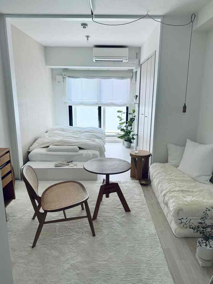 白とベージュの落ち着いた色調のワンルーム寝室兼リビングルーム、ナチュラルな木製家具と白いカーペットが配置された雰囲気のあるインテリア