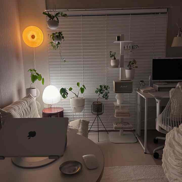 White and beige toned living room featuring numerous plants and standing lamps, creating a warm and cozy home office atmosphere