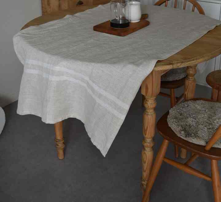 ベージュのリネンのテーブルクロスが掛けられた丸い木製のダイニングテーブルと、クッションのある木製チェアが並ぶナチュラルなダイニング空間