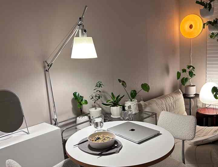 柔らかなベージュとホワイトの色調のダイニングルーム、丸テーブルとアルテミデのテーブルランプ、緑の植物が調和した落ち着いたホームオフィス空間