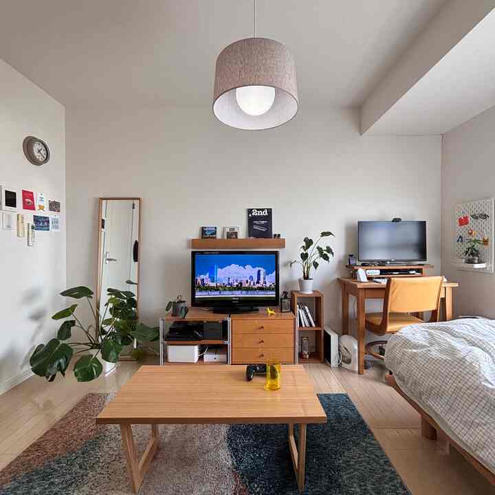 ナチュラルな木目調と白い壁の1Kワンルーム、小さな机とベッド、観葉植物が配置された落ち着いた一人暮らしの空間