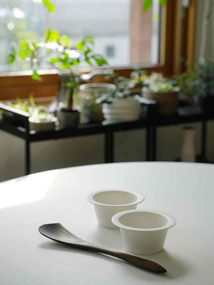 明るい自然光が差し込む窓辺に植物と木目調の調理器具が置かれたナチュラルな白いダイニングテーブル