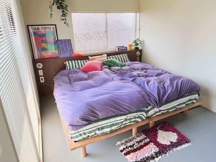 ライトグレーの床と木目調のベッドフレームがある寝室、紫の掛け布団カバーとカラフルなクッションが配置された雰囲気のある空間