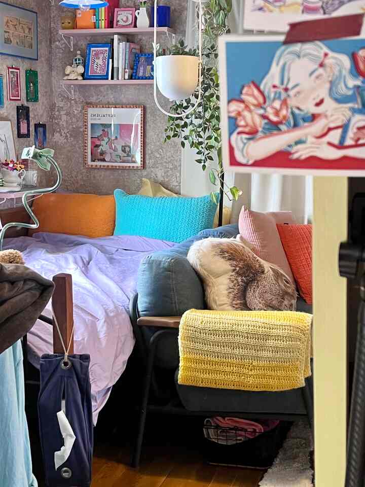 A colorful bedroom featuring cushions in vivid shades and lavender duvet cover with concrete patterned wallpaper creating a cozy atmosphere