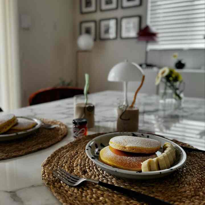 白とナチュラルな色調のダイニングルーム、IKEAの皿にパンケーキがのり、テーブルランプが置かれた雰囲気のあるホームカフェ空間