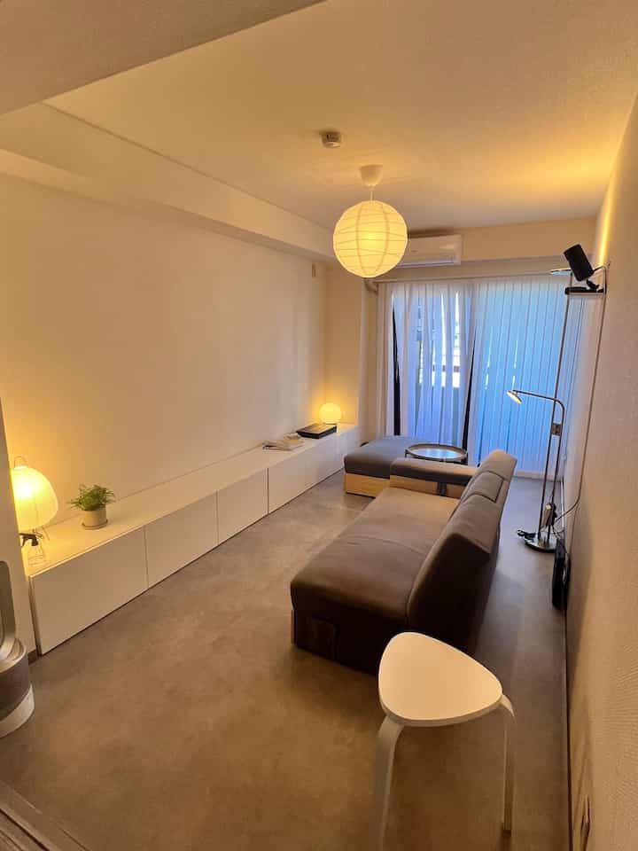 Narrow living room with white walls and long storage cabinet, brown sofa and white stool, pendant light creating a soft natural modern atmosphere