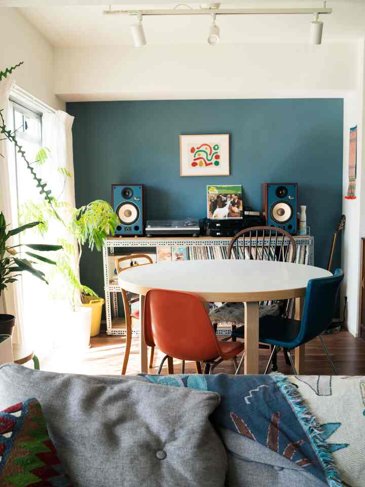 青いアクセントウォールと植物が配置された北欧風のダイニングルーム、中央に丸テーブルとカラフルな椅子がある印象的な空間