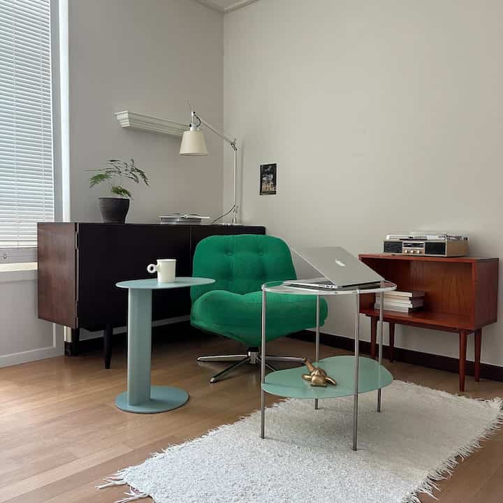 明るいナチュラルトーンの書斎空間、緑のアームチェアとミント色のテーブル、木製収納棚と照明が配置された落ち着いたインテリア