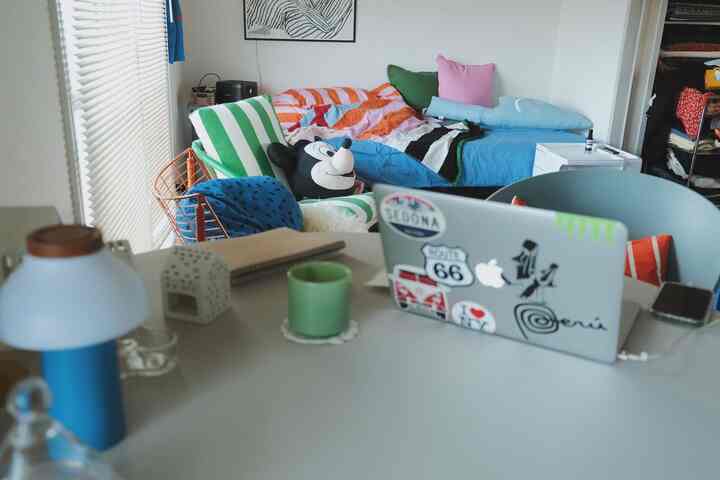 Bright monotone living-bedroom space featuring white desk with laptop and blue table lamp in a compact room
