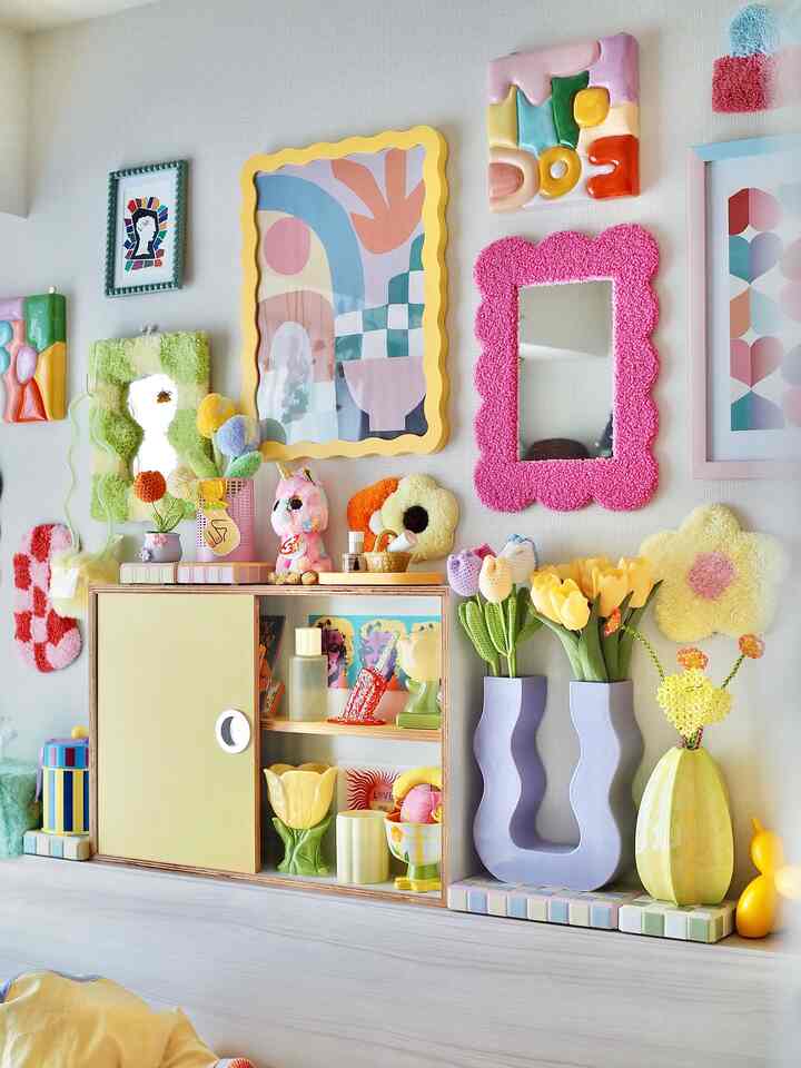 A yellow and pastel-toned bedroom wall featuring colorful art pieces, decorative vases, and mirrors in a stylish arrangement