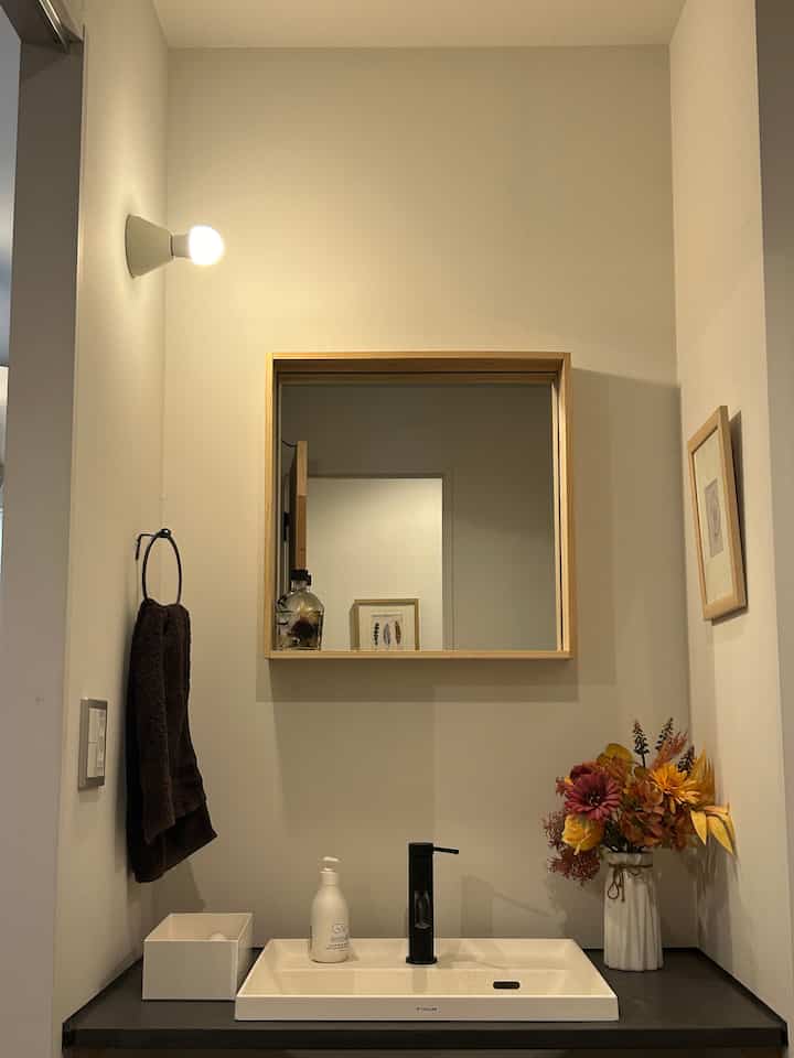 白と黒のトーンのバスルーム洗面台、中央に木製の鏡があり、右側に華やかな造花の花瓶が置かれた落ち着いたインテリア