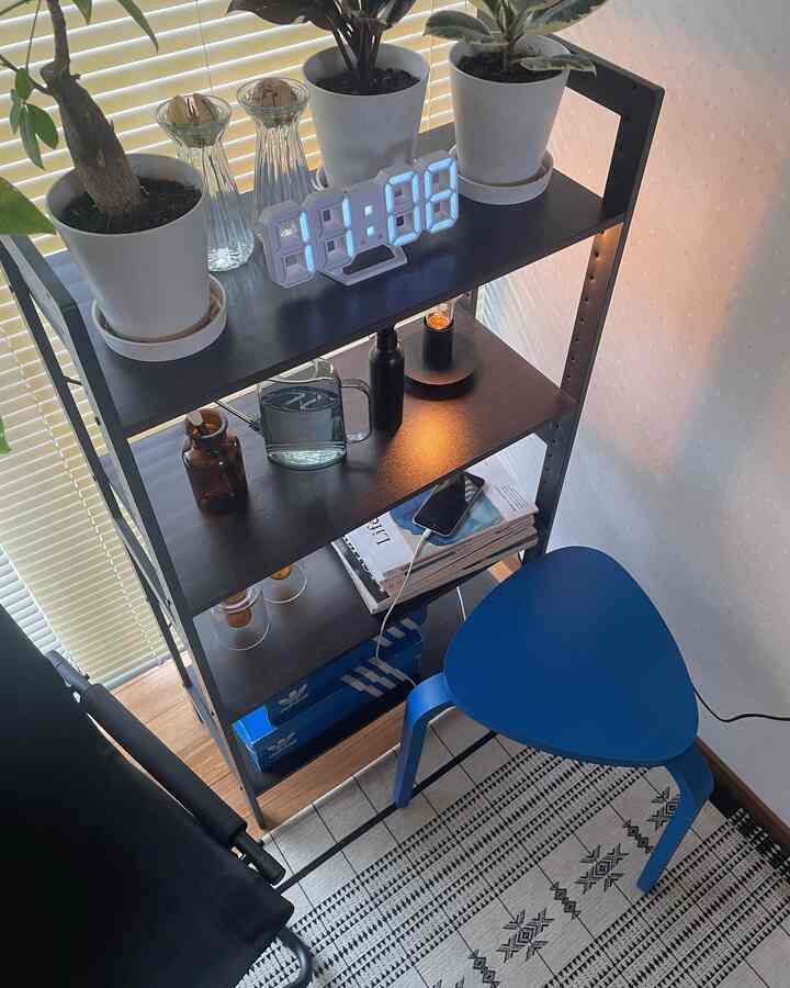 Bright living room corner featuring a blue stool and black metal shelf with plants and digital clock, showcasing neat storage and decor