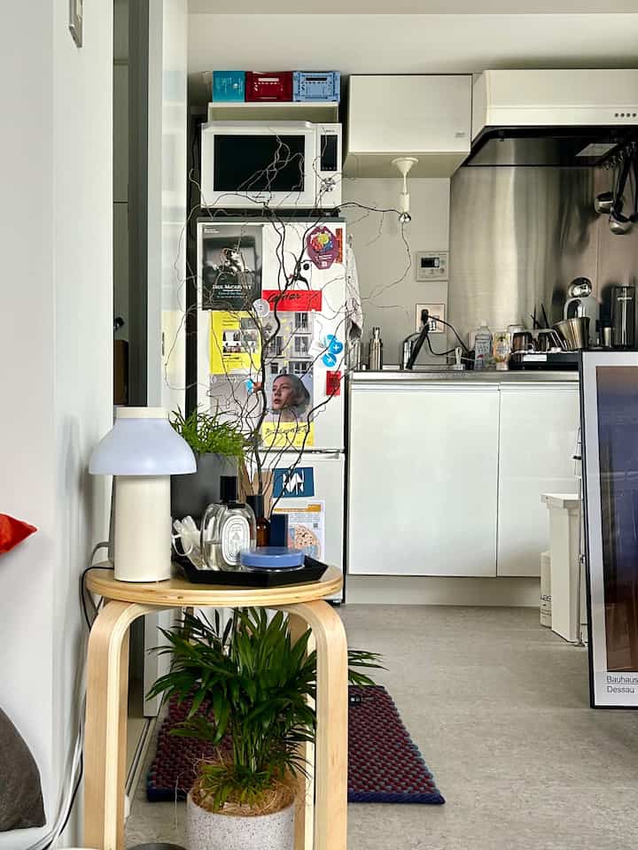 白と木目調のワンルームキッチン、素朴なサイドテーブルとスツール、植物、テーブルランプが配置された落ち着いた雰囲気のインテリア