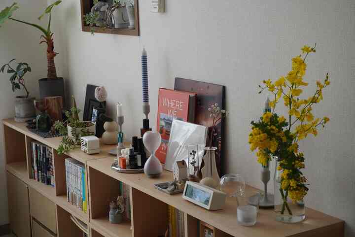 Natural wood tone bookshelf against a white wall in a living room display corner, featuring yellow flowers and various decorative items creating a cozy atmosphere