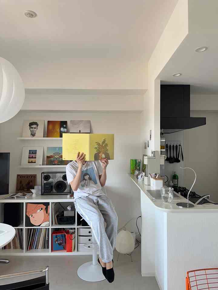 ホワイトトーンのワンルームキッチンとリビングスペース、ミニマルな家具と本棚、中央のスツールに座る人物がいるモダンなインテリア