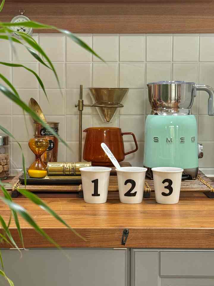 温かみのあるブラウン木目調キッチン、ミント色のSMEGミルクフォーマーと個性的な数字入り陶器のカップが置かれた雰囲気のあるおうちカフェ空間