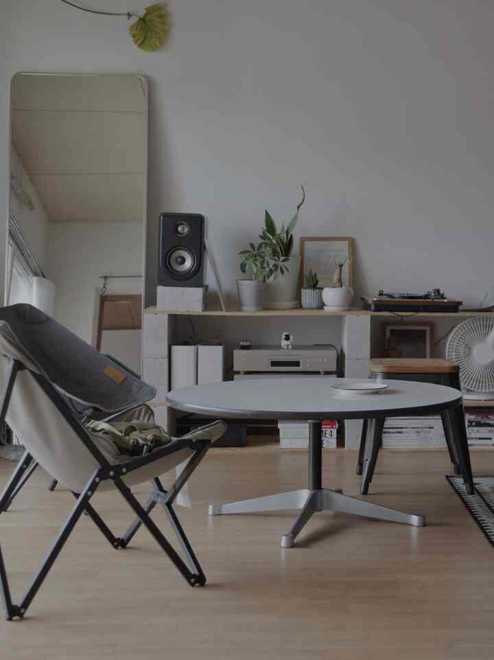 白壁と茶色の床のワンルームリビング、ラウンドローテーブルとアウトドアチェア、植物とスピーカーが置かれたミニマルでナチュラルな雰囲気のインテリア