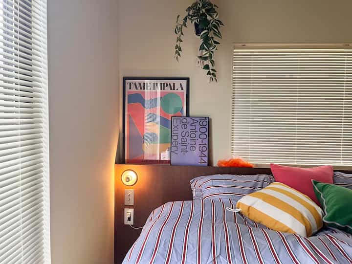 ホワイトとブラウンの色調の寝室、カラフルなポスターとストライプのベッドカバー、クッションが配置されたシンプルで落ち着いた印象のインテリア