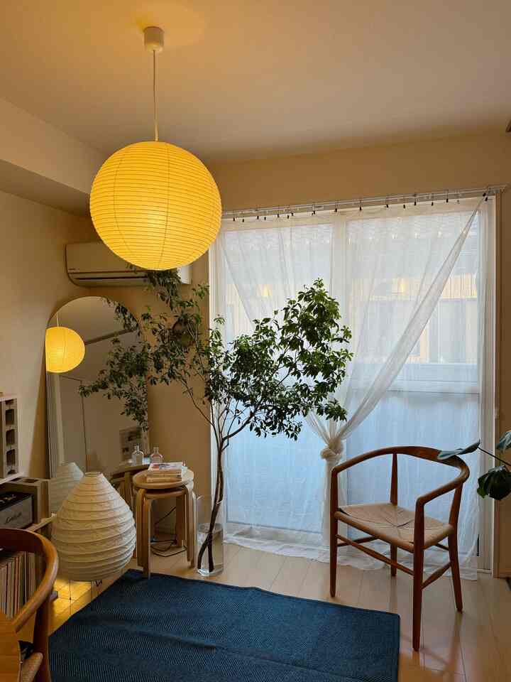 ナチュラルな木目調と白を基調としたリビング、アームチェアと大きな植物、ラグ、イサム・ノグチ風の照明が配置された印象的な空間