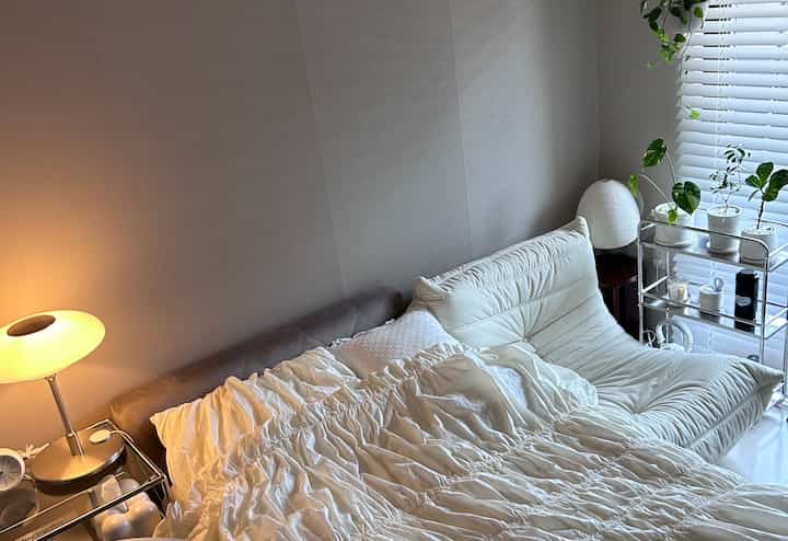 Cozy bedroom with soft brown walls and white bedding, featuring a bed, table lamp, and plants for a natural ambiance