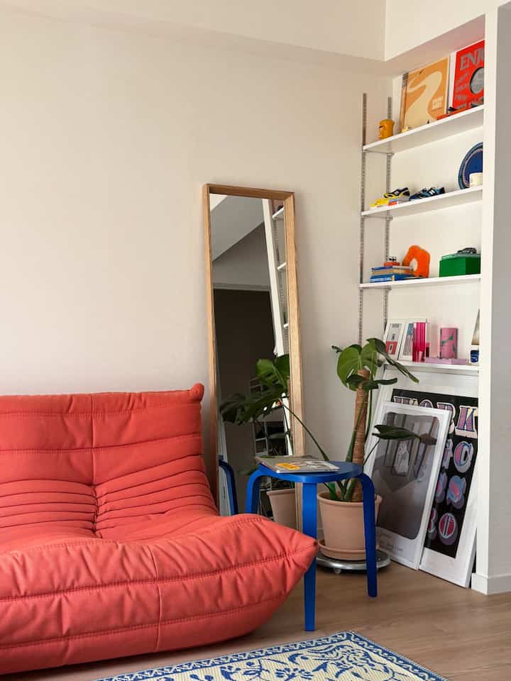 Natural toned living room featuring a vibrant coral sofa, blue stool, large wooden-framed mirror, and green plant for a clean look