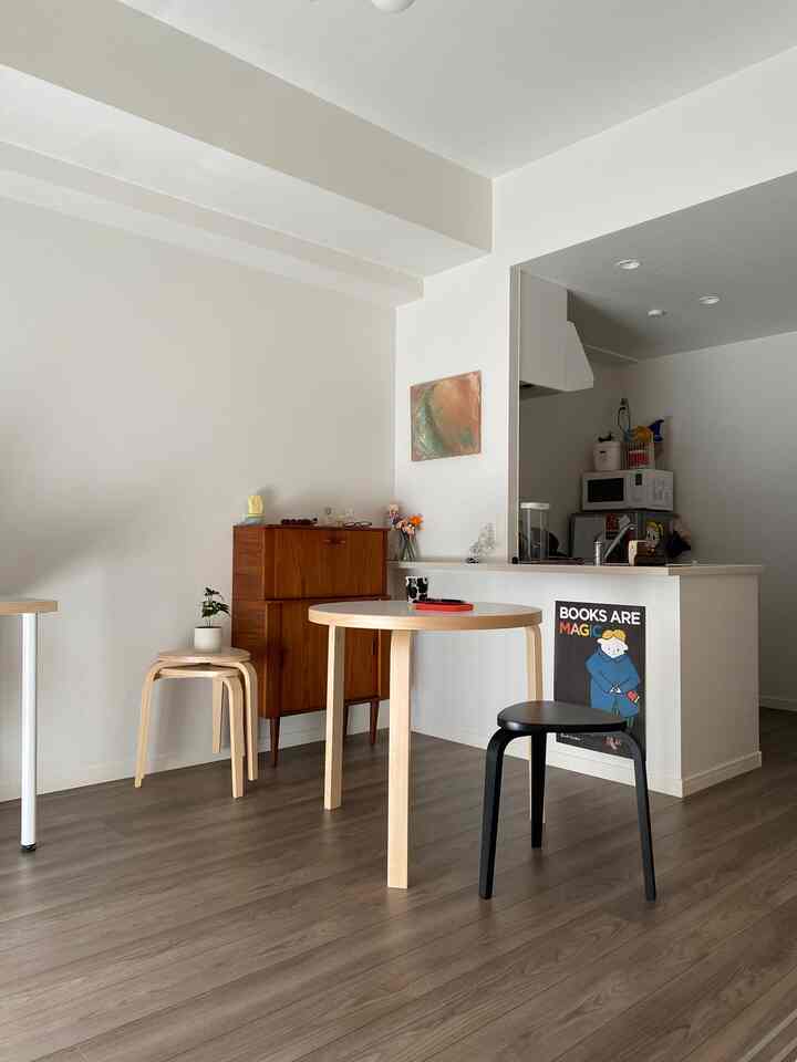 白い壁と天井のナチュラルスタイルリビング、丸い木目調ダイニングテーブルとスツールが配置されたすっきりとした空間