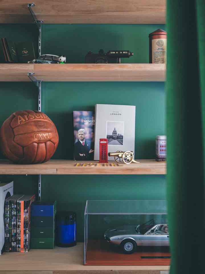 ナチュラルな木目調と緑の壁の書斎、棚にはミニカーやヴィンテージのサッカーボール等のコレクティブルが並ぶ雰囲気あるインテリア