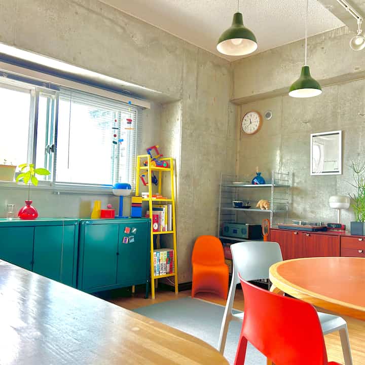 コンクリート打ちっぱなしの壁と木製円形テーブル、多色のプラスチックチェアが配置されたワンルームのダイニングスペース